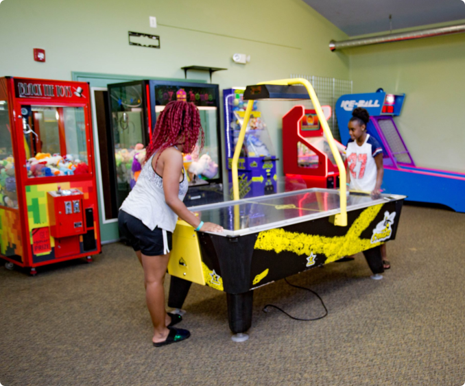 Two people playing air hockey in an arcade, surrounded by various game machines and claw cranes.