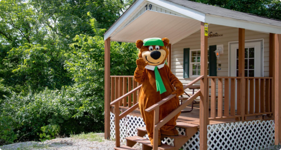 A bear mascot stands on the porch of a rustic cabin