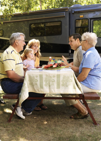 Five people, including a young child, sit at a picnic table with food outdoors near a large RV, engaging in conversation.